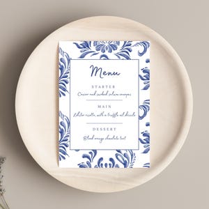 Może przedstawiać: Biała karta menu z niebieskim kwiatowym wzorem na obrzeżach. Menu zawiera następujące pozycje: Przystawka - Kawior i wędzone kanapki z łososiem, Danie główne - Risotto z homara z polewą z truflowego oleju, Deser - Tarta czekoladowa z krwawymi pomarańczami.
