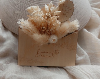 May include: A beige wooden envelope with dried flowers and the text "Bonne fête Mamie". A small wooden tag with the names "Emma Louis Mila" is attached. A textured, off-white vase is in the background.