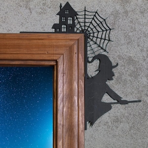 May include: A wooden framed picture with a night sky scene. A black silhouette of a witch on a broom, a haunted house, and a spiderweb are attached to the frame, creating a Halloween theme.