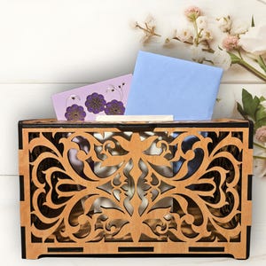 May include: A wooden decorative box with a cutout design featuring intricate floral patterns. The box is brown with black accents and has a light wood interior. Two cards are visible inside the box, one light blue and one light purple with a floral design.