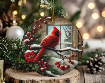 May include: Round Christmas ornament featuring a vibrant red cardinal perched on a snow-covered branch with red berries. The ornament has a winter scene with a window in the background. The ornament is hanging from a gold string.