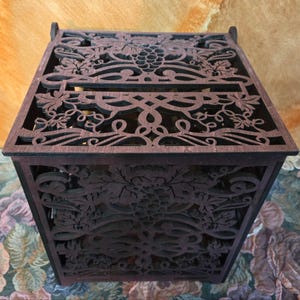 May include: A dark brown wooden box with intricate laser-cut grape vine and leaf designs. The box has a rectangular shape with a slot on top and is likely used for storage or decoration. The box is made of wood and has a detailed, decorative pattern.
