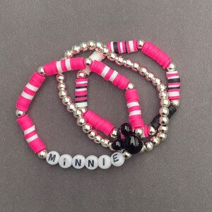 May include: Three pink and white striped bracelets with silver beads and a black Mickey Mouse charm. The bracelets spell out the name "MINNIE" in white letters.