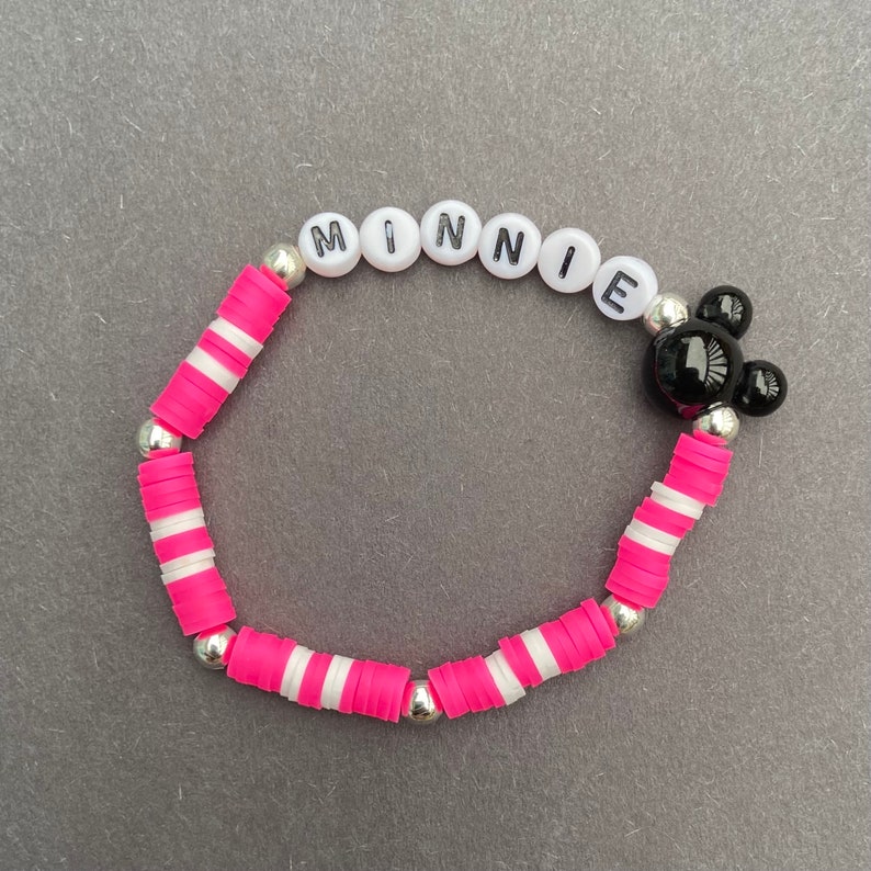 Puede incluir: Una pulsera de rayas rosa y blancas con la palabra "MINNIE" escrita en letras negras sobre cuentas blancas. La pulsera tambi&eacute;n tiene un dije de cabeza de Mickey Mouse en blanco y negro.