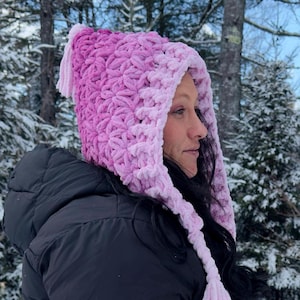 May include: A handmade, crochet hood in shades of pink and lavender. The hood features a textured stitch pattern and long tassels. The person is wearing a black puffer jacket in a snowy, outdoor setting.