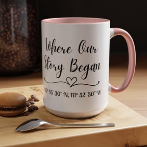 May include: White ceramic mug with a pink interior and handle. The mug features the text "Where Our Story Began" in black script, along with a heart symbol and coordinates. A macaron and spoon are on a wooden surface.