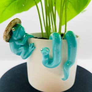 May include: A white cylindrical plant pot featuring a teal frog figurine wearing a small straw hat and a teal snake figurine attached to the rim. The frog and snake have small, reflective, jewel-like eyes. A green leafy plant grows inside the pot.