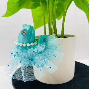 May include: A turquoise chameleon figurine wearing a white pearl necklace and a light blue tulle tutu skirt decorated with small blue star and circle sequins. The chameleon has a single visible pink eye and is positioned in front of a green plant.