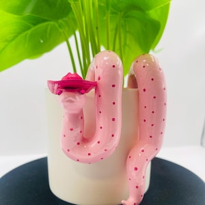 May include: A light pink snake figurine with darker pink polka dots, wearing a small, bright pink cowboy hat. The snake is coiled around the exterior of a plain, cylindrical, off-white plant pot, with its head positioned near the top rim.