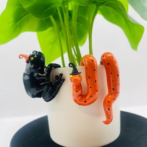 May include: A glossy black frog figurine wearing a small orange hat and a glossy orange snake figurine with black spots wearing a small black hat, both perched on the rim of a plain white cylindrical plant pot containing a green plant.