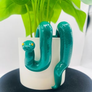 May include: A white cylindrical plant pot featuring a glossy, dark teal snake sculpture wrapped around the exterior. The snake has two small, bright yellow circular eyes. A green leafy plant is positioned inside the white container.