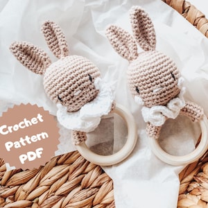 May include: Two crocheted bunny rattle toys with white ruffled collars attached to wooden rings. The toys are on a white tissue paper background with a brown wicker basket.