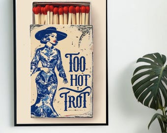 May include: A framed art print featuring a vintage matchbox design. The matchbox illustration depicts a woman in a blue dress and hat, with the words "Too Hot Trot" in blue text. The matchbox contains red-tipped matches.