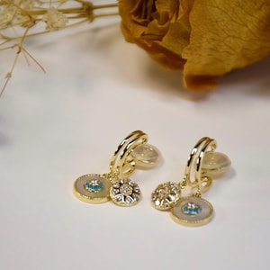 Peut inclure: Une paire de boucles d'oreilles créoles dorées avec un pendentif représentant un œil maléfique blanc et bleu et un plus petit pendentif avec des strass transparents.