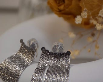 May include: Silver-coloured dangle earrings with a cascading chain design. The earrings are displayed on a white surface, with a blurred background featuring a dried orange rose and small white flowers. The earrings have a post-back closure.
