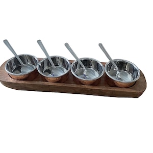 May include: A set of four small copper bowls with stainless steel spoons, arranged on a wooden tray. The bowls are ideal for serving small portions of food or condiments.