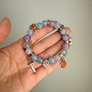 May include: A beaded bracelet featuring translucent light blue and purple crackle glass beads interspersed with gold-toned metal spacers. The bracelet includes several small metal charms, including a sword, a shield, a triangular emblem, and a mask.
