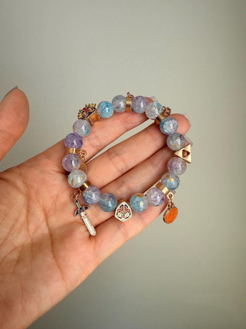 May include: A beaded bracelet featuring translucent light blue and purple crackle glass beads interspersed with gold-toned metal spacers. The bracelet includes several small metal charms, including a sword, a shield, a triangular emblem, and a mask.