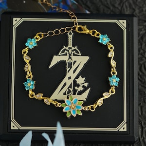 Op de afbeelding: Een goudkleurige armband met blauwe bloemaccenten en een bladerontwerp. De armband wordt weergegeven op een zwart vierkant met een goudkleurig embleem. Het embleem toont een zwaard en de letter Z.