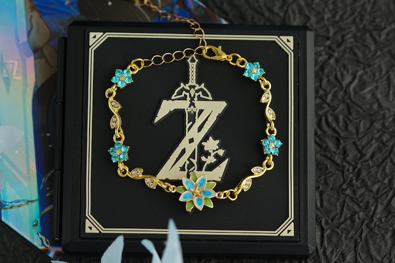 Op de afbeelding: Een goudkleurige armband met blauwe bloemaccenten en een bladerontwerp. De armband wordt weergegeven op een zwart vierkant met een goudkleurig embleem. Het embleem toont een zwaard en de letter Z.
