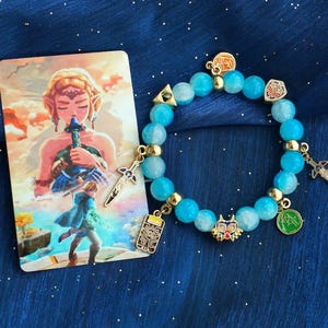 May include: A blue beaded bracelet with gold accents and charms, including a sword, a shield, and a Triforce symbol. A card with a watercolor illustration of a character from a video game is next to the bracelet.