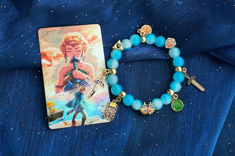 May include: A beaded bracelet featuring light blue translucent beads, gold-toned spacers, and various metal charms including swords, a mask, and a shield. Beside the bracelet is a rectangular card depicting a character holding a sword.