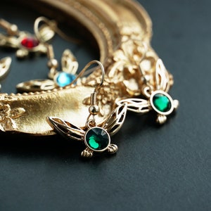 May include: Gold-coloured earrings featuring a leaf design and a round, emerald-green gemstone. The earrings are displayed on a gold decorative frame. Other earrings with red and blue gemstones are visible.