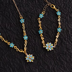 Op de afbeelding: Een goudkleurige ketting- en armbandset. De sieraden hebben een bloemenontwerp met blauwe en groene emaille accenten. De ketting heeft een hanger met een grote bloem en de armband heeft een bijpassend ontwerp. De set staat op een zwarte achtergrond.
