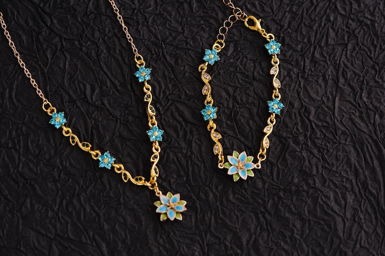 Op de afbeelding: Een goudkleurige ketting- en armbandset. De sieraden hebben een bloemenontwerp met blauwe en groene emaille accenten. De ketting heeft een hanger met een grote bloem en de armband heeft een bijpassend ontwerp. De set staat op een zwarte achtergrond.