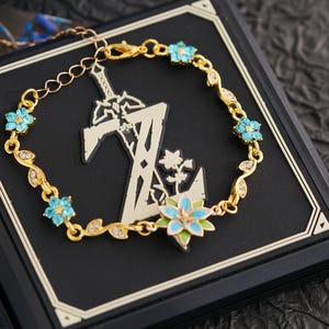 Op de afbeelding: Goudkleurige armband met een bloemmotief en lichtblauwe accenten. De armband is versierd met kleine, sprankelende kristallen en blauwe bloemvormige versieringen. De armband wordt gepresenteerd op een zwarte ondergrond.