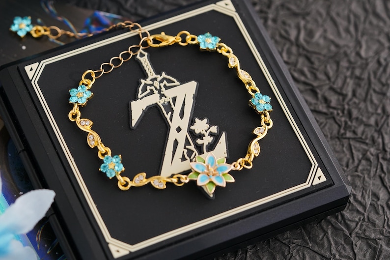 Op de afbeelding: Goudkleurige armband met een bloemmotief en lichtblauwe accenten. De armband is versierd met kleine, sprankelende kristallen en blauwe bloemvormige versieringen. De armband wordt gepresenteerd op een zwarte ondergrond.