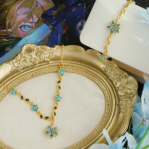 Op de afbeelding: Een goudkleurige bloemen haarband en bijpassende armband met lichtblauwe en groene accenten. De haarband wordt getoond op een gouden sierlijke lijst. Lichtblauwe bloemen zijn op de achtergrond.