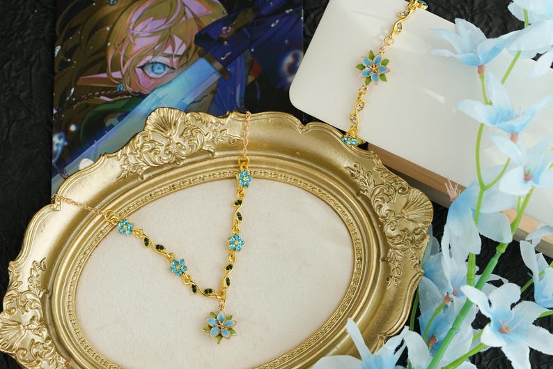 Op de afbeelding: Een goudkleurige bloemen haarband en bijpassende armband met lichtblauwe en groene accenten. De haarband wordt getoond op een gouden sierlijke lijst. Lichtblauwe bloemen zijn op de achtergrond.
