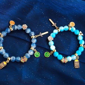 May include: Two beaded bracelets featuring round blue stones and gold-toned metal charms. The charms include miniature swords, shields, triangular symbols, circular icons, and a colorful mask. The beads on the left are marbled blue, while the beads on the right are a solid, bright sky blue.