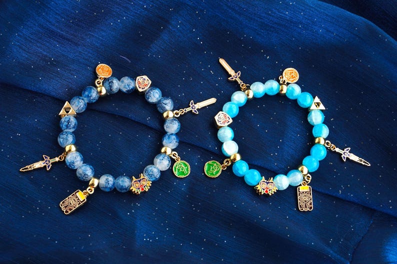 May include: Two beaded bracelets featuring round blue stones and gold-toned metal charms. The charms include miniature swords, shields, triangular symbols, circular icons, and a colorful mask. The beads on the left are marbled blue, while the beads on the right are a solid, bright sky blue.