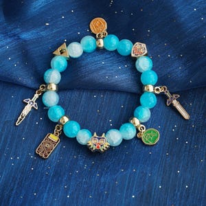 May include: A stretch bracelet featuring round, light blue, semi-translucent beads interspersed with gold-toned metal spacers. The bracelet includes several hanging charms, including a colorful mask, two swords, a shield, a triangle, and a rectangular tablet.