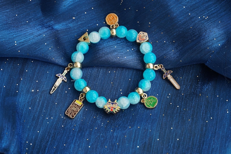 May include: A stretch bracelet featuring round, light blue, semi-translucent beads interspersed with gold-toned metal spacers. The bracelet includes several hanging charms, including a colorful mask, two swords, a shield, a triangle, and a rectangular tablet.