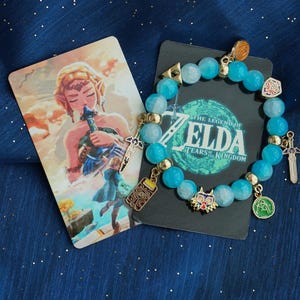 May include: A beaded bracelet featuring light blue translucent beads and gold-toned metal charms, including a sword, a mask, and a rectangular device. The bracelet is displayed on a black card with The Legend of Zelda Tears of the Kingdom text.