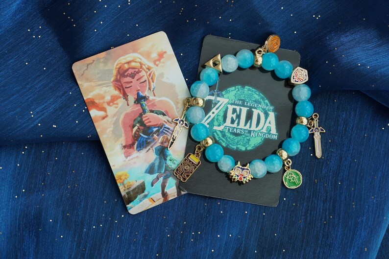 May include: A beaded bracelet featuring light blue translucent beads and gold-toned metal charms, including a sword, a mask, and a rectangular device. The bracelet is displayed on a black card with The Legend of Zelda Tears of the Kingdom text.