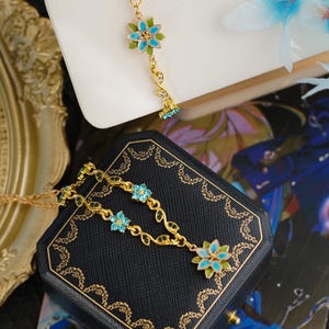 Op de afbeelding: Goudkleurige sieradenset met een armband en ketting. Beide stukken tonen blauwe en groene bloemmotieven met delicate bladaccenten. De sieraden worden gepresenteerd op een wit oppervlak en een donkerblauwe sieradendoos.