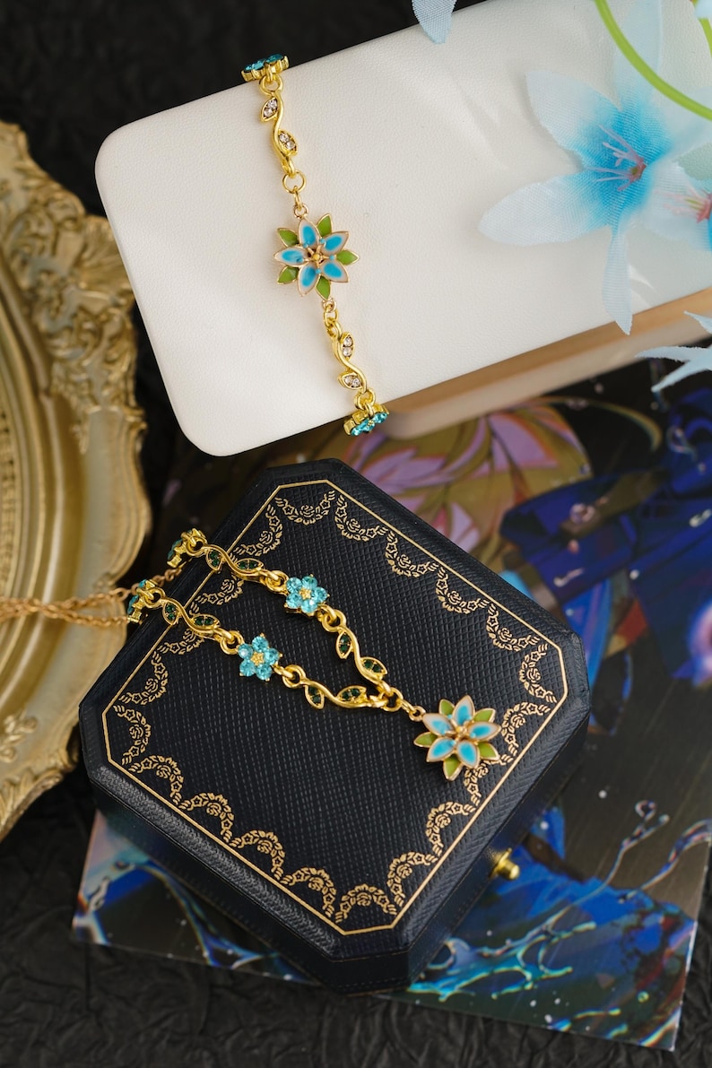 Op de afbeelding: Goudkleurige sieradenset met een armband en ketting. Beide stukken tonen blauwe en groene bloemmotieven met delicate bladaccenten. De sieraden worden gepresenteerd op een wit oppervlak en een donkerblauwe sieradendoos.