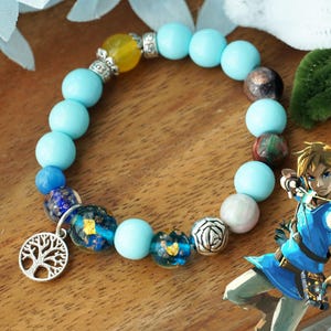 Puede incluir: Un brazalete azul y plateado con un dije de árbol de la vida. El brazalete tiene cuentas azules y plateadas, una cuenta amarilla y una cuenta de vidrio con destellos dorados. El brazalete está sobre una superficie de madera.