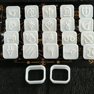 May include: A set of white, square game pieces featuring different symbols, displayed on a black surface with gold detailing. Two white square frames are in the foreground. The pieces are likely related to a game or fantasy theme.