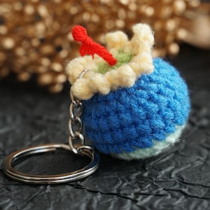 May include: A blue, green, and yellow crocheted keychain shaped like a small ball with a red crocheted stem on top. The keychain is attached to a silver metal key ring.