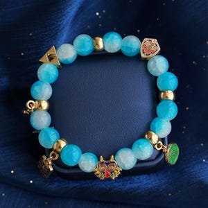 May include: A beaded bracelet featuring round, translucent light blue and white marbled beads interspersed with gold-toned metal spacers. The bracelet includes several small metal charms, including a colorful mask, a shield, and a triangular emblem.