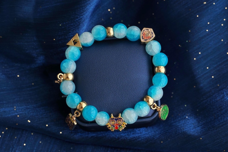May include: A beaded bracelet featuring round, translucent light blue and white marbled beads interspersed with gold-toned metal spacers. The bracelet includes several small metal charms, including a colorful mask, a shield, and a triangular emblem.