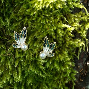May include: A pair of gold earrings with a white and blue enamel design, featuring a pearl accent. The earrings are shaped like leaves and are set against a green mossy background.