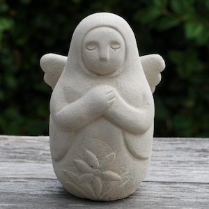 May include: A light beige stone sculpture of an angel with closed eyes and hands clasped over the chest. The figure features small wings on the sides and a carved flower design at the base, with a smooth, matte texture throughout the entire piece.