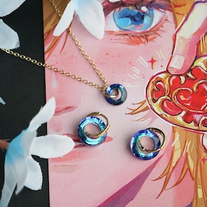 Op de afbeelding: Een gouden ketting met een blauw-gouden hanger en twee bijpassende blauw-gouden ringen. De sieraden worden gepresenteerd op een roze achtergrond met een cartoonillustratie van een hartvormige hanger en een vrouwenoog.