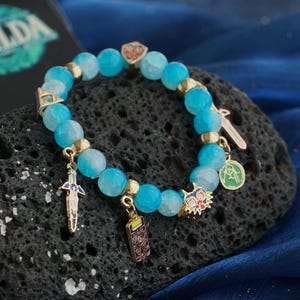 May include: A beaded bracelet featuring faceted light blue stones, gold-toned spacers, and various metal charms including a sword, a rectangular emblem, a colorful mask, and a circular green pendant, with a Zelda logo visible in the background.